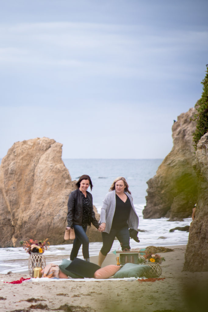Two women smile while holding hands as they walk up to a picnic setup on El Matador State Beach in Malibu, California.